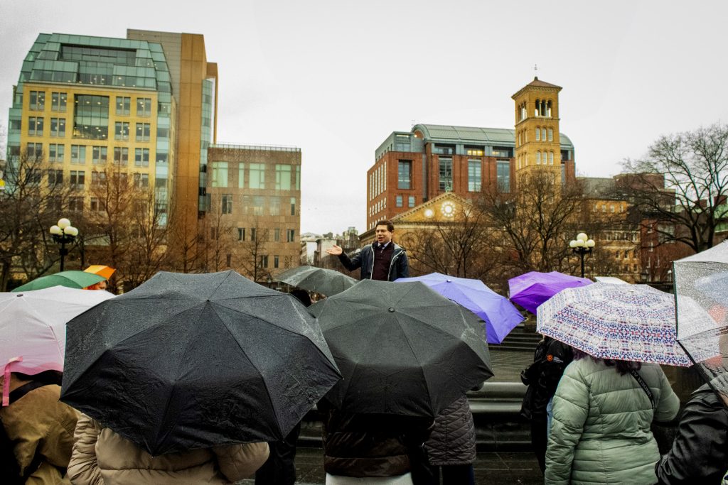 Ulysses Butler addresses students at the NYU campus on a rainy day. The students are facing him holding umbrellas. 